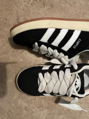 Svarta Adidas Campus sneakers - Säljer ett par svarta Adidas Campus sneakers med klassiska vita ränder på sidorna och breda vita skosnören. Skorna har en retro look med låg profil och gummisula i brunt. Ovandelen är i mocka och textil, perfekt för dig som gillar streetwear.