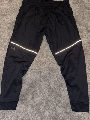Svarta träningsbyxor Under Armour - Snygga svarta träningsbyxor från Under Armour med reflekterande detaljer över låren och logga på vänster ben. Byxorna har resår i midjan, smal passform och muddar vid bensluten. Perfekta för löpning eller gym. Tillverkade i ett lätt och snabbtorkande material.