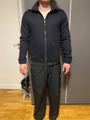 Mörkblå cardigan från Massimodutti  - Snygg mörkblå jacka från Massimodutti med dragkedja och huva. Jackan har två sidofickor och detaljer i grått vid krage och nederkant. Perfekt för dig som vill ha en stilren och enkel jacka till vardags. Materialet är mjukt och jackan har en modern passform.
