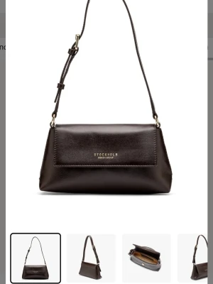 Brun axelväska från Stockholm Design Group - Snygg mörkbrun axelväska i skinn från Stockholm Design Group. Väskan har justerbar axelrem, guldfärgade detaljer och ett stilrent, minimalistiskt uttryck. Perfekt storlek för det viktigaste och med logga framtill.