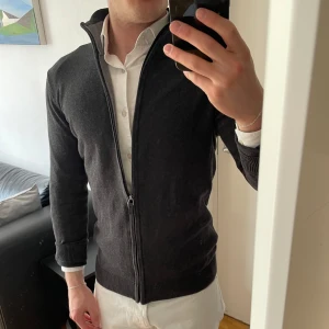 Mörkgrå kofta från Tom Tailor - Säljer en stilren mörkgrå kofta från Tom Tailor med hel dragkedja och hög krage. Tröjan är stickad i mjukt material och har långa ärmar samt ribbade muddar. Perfekt för lager-på-lager och funkar till många olika outfits. Bra skick, en gnutta urtvättad men märks inte av.