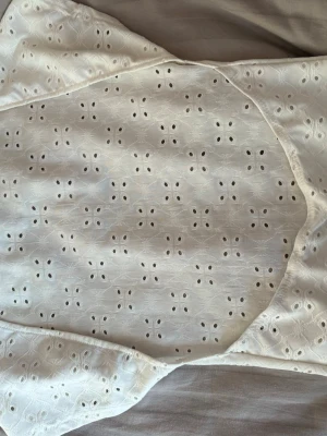 Vit broderad blus med hålmönster - Söt vit blus med korta ärmar och broderade hålmönster över hela plagget. Toppen har en feminin och luftig känsla, perfekt för varma dagar. Materialet känns mjukt och lätt, och den har en klassisk passform som passar till mycket.