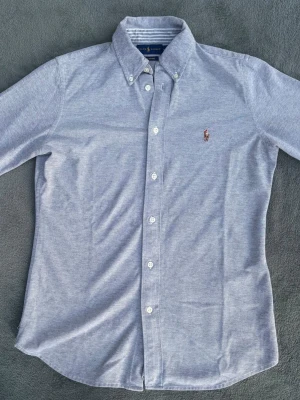 Ljusgrå skjorta från Polo Ralph Lauren - Klassisk ljusgrå skjorta från Polo Ralph Lauren med broderad logga på bröstet. Skjortan har lång ärm, knappar hela vägen och button-down krage. Tillverkad i mjuk bomull för en bekväm känsla och stilren look.