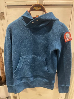 Blå hoodie från Fils Kolm med patch - Parajumpers Mysig blå hoodie med stor känguruficka och ribbade muddar. Huvan har en cool knappdetalj och insidan är röd. På ärmen sitter en broderad patch med text och motiv. Perfekt för dig som gillar streetwear och vill ha något unikt i garderoben.