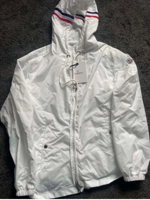 Vit vindjacka från Moncler - Snygg vit vindjacka från Moncler med huva och dragkedja framtill. Jackan har Moncler-logga på ärmen, två fickor med knappar och elastiska muddar. Huvan har röda och blå ränder som detalj samt dragsko för justering. Perfekt för en sportig och trendig look.