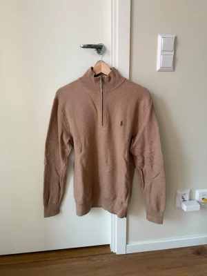 Halvzip  Polo Ralph Lauren - Snygg beige stickad tröja från Polo Ralph Lauren med halvzip och hög krage. Tröjan har lång ärm och den klassiska lilla loggan broderad på bröstet. Perfekt för lager på lager och enkel att matcha till olika outfits. Passsar S/M