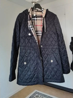 Svart  vindjacka Burberry - Snygg svart  quiltad vindjacka från Burberry med silverfärgade knappar och dragsko i huvan. Jackan har ett klassiskt rutmönster i tyget och detaljerade knappar med Burberry-logga. Perfekt för dig som vill ha en stilren och tidlös look. Fattas en knapp bara 