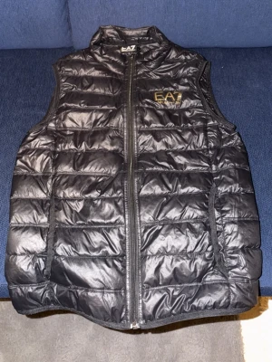 Svart dunväst från EA7 Emporio Armani - Snygg svart dunväst från EA7 Emporio Armani med guldfärgad logga på bröstet. Västen har quiltad design, hög krage och dragkedja framtill. Perfekt att slänga över en hoodie för en cool och sportig look.
