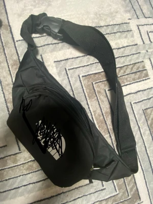 Svart magväska från The North Face - Snygg svart magväska från The North Face med justerbart band och plastspänne. Väskan har en stor dragkedjeförsedd ficka och är tillverkad i slitstarkt tyg. Perfekt för att bära det viktigaste när du är på språng.