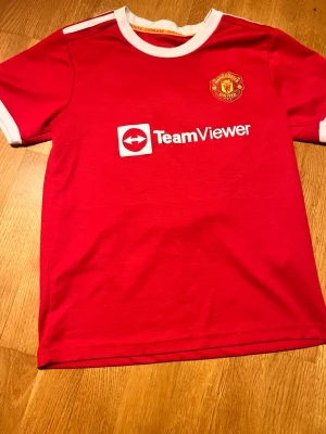 Manchester United matchtröja röd - Röd Manchester United matchtröja med vitt TeamViewer-tryck på bröstet och klubbmärke på vänster sida. Tröjan har vita detaljer vid krage och ärmslut. Perfekt för fotbollsträning eller att bära på matchdag. Tillverkad i lätt och ventilerande material.