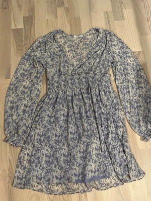 Blommig omlottklänning från NA-KD - Söt kort omlottklänning från NA-KD med blommigt mönster i blått och beige. Klänningen har långärmade ärmar, v-ringning och smockad midja som ger en fin passform. Perfekt för vår och sommar!