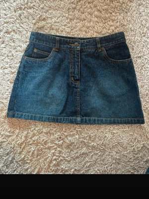 Mörkblå jeanskjol -  mörkblå jeanskjol med fem fickor och knappstängning framtill. Kjolen har en rak och kort passform, perfekt för att styla med sneakers eller boots.