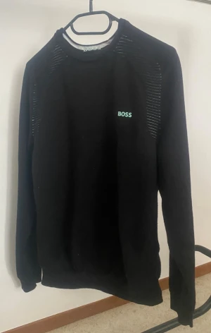 Svart sweatshirt från BOSS - Svart sweatshirt från BOSS med diskret logga i grönt på bröstet. Tröjan har rund halsringning, långa ärmar och detaljer med tunna ränder vid axlar och övre rygg. Perfekt för en stilren och avslappnad look.