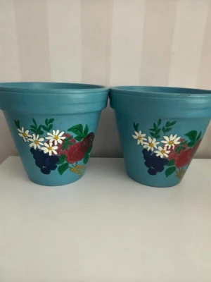 Blå handmålade blomkrukor - Två blå blomkrukor i keramik med handmålade blommor i vitt, rött och blått. Krukorna har en klassisk konisk form och dekoren består av prästkragar, röda rosor och blå bär. Perfekta för att ge lite färg till fönsterbrädan eller balkongen. .Mått diameter 15 cm, höjd 14 cm.