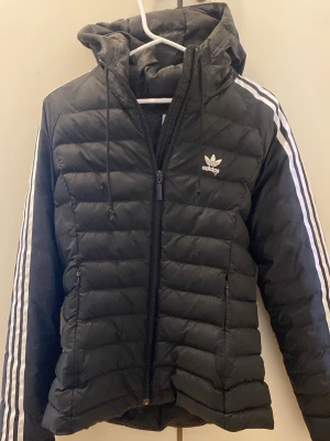 Svart dunjacka med huva från Adidas - Snygg svart dunjacka från Adidas med klassiska vita ränder längs ärmarna och Adidas-logga på bröstet. Jackan har huva, dragkedja framtill och två sidofickor med dragkedja. Perfekt för kalla vinterdagar och sportig streetstyle.