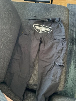 Svarta cargobyxor från Corteiz  - Säljer ett par svarta cargobyxor från Corteiz med flera stora fickor och en cool vit tryckt grafik framtill. Byxorna har justerbart bälte i midjan och en gul patch på sidan. Perfekta för dig som gillar streetwear och funktionella detaljer.