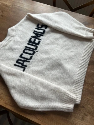 Vit stickad tröja från Jacquemus - Säljer en vit stickad tröja från Jacquemus med svart logotyp i grafisk stil över ryggen. Tröjan har rund halsringning, långa ärmar och ribbade muddar vid ärmslut och nederkant. Perfekt för dig som gillar trendiga och stilrena plagg. Hör av er vid minsta fundering 😁 Storlek S-M