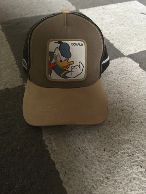 Brun keps med Kalle Anka från Goorin Bros - Säljer en brun och beige keps från Goorin Bros med ett stort Kalle Anka-märke framtill. Kepsen har svart mesh baktill och justerbar snapback. Materialet är en mix av bomull och polyester. Perfekt för dig som gillar Disney och vill sticka ut med en unik accessoar.