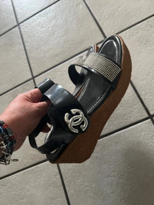 Svarta platåsandaler storlek 37 - Snygga svarta platåsandaler med bred rem i lackmaterial och silverfärgad logga på sidan. Främre remmen är dekorerad med glittrande stenar och sulan är i korkliknande material för en trendig look.