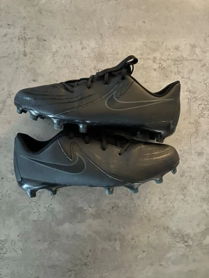 Svarta Nike fotbollsskor - Snygga helsvarta fotbollsskor från Nike med diskret swoosh-logga på sidan. Skorna har klassisk låg profil, snörning och fasta dobbar för spel på gräs. Tillverkade i slitstarkt syntetmaterial som ger bra bollkontroll och komfort på planen.