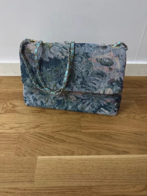 Blommig handväska med kedjerem - Unik handväska i gobelängtyg med blommigt mönster i blå, grå och gröna toner. Väskan har en flätad kedjerem i guld och blått, samt en mjuk, rektangulär form. Perfekt för dig som gillar vintage vibes och vill sticka ut.