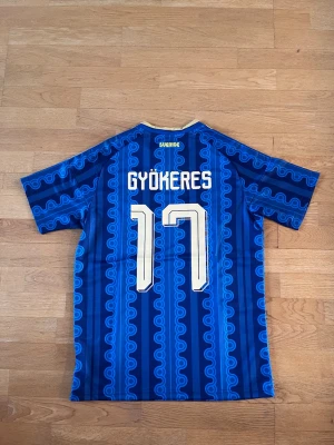 Sverige fotbollströja Gyökeres #17 - Blå Sverige fotbollströja från Adidas med Gyökeres och nummer 17 på ryggen. Tröjan har guldiga detaljer, klassiska Adidas-ränder på axlarna och Sveriges landslagsemblem på bröstet.