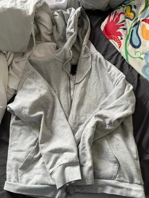 Grå hoodie med dragkedja från 157 - Säljer en grå hoodie från 157 med dragkedja framtill och stor huva. Hoodien har fickor på sidorna och är gjord i mjukt, fleecefodrat material som känns riktigt skönt mot huden. Perfekt för chill dagar eller när du vill ha något bekvämt och stilrent.