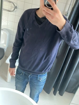 Mörkblå v-ringad tröja - Mörkblå v-ringad tröja med långa ärmar och enkel design. Tröjan har en liten broderad logga på bröstet och är tillverkad i ett mjukt material som passar perfekt till jeans. Perfekt för en avslappnad stil.