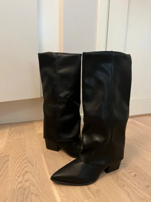 Svarta knähöga boots med spetsig tå - Står strl 40 men väldigt små i strl är mer som strl 39!   Säljer ett par svarta knähöga boots i slätt konstläder med spetsig tå och låg klack. Stövlarna har en stilren design och passar perfekt till både jeans och kjol. De har en något lösare passform upptill och ger en trendig siluett. Lite missformad på vänster sko se bild.
