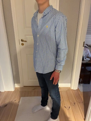 Blårutig skjorta från Polo Ralph Lauren - Snygg blårutig skjorta från Polo Ralph Lauren i slim fit-modell. Skjortan har klassisk krage, långa ärmar och knappar framtill. Tillverkad i mjuk bomull och har den ikoniska gula loggan broderad på bröstet. Perfekt för en clean och stilren look. Tröjan är i nyskick, dvs. 10/10. 