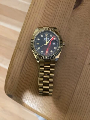 Guldfärgad klocka med svart urtavla - Snygg guldfärgad klocka med svart urtavla och guldfärgade detaljer. Urtavlan har datumvisning och markeringar med stenar. Länken är i metall och ger en lyxig känsla. Perfekt accessoar för dig som vill sticka ut.