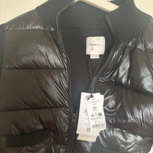 Svart pufferjacka från Gina Tricot - Snygg svart pufferjacka från Gina Tricot med glansig yta och ribbstickade ärmar samt krage. Jackan har två framfickor och dragkedja framtill. 