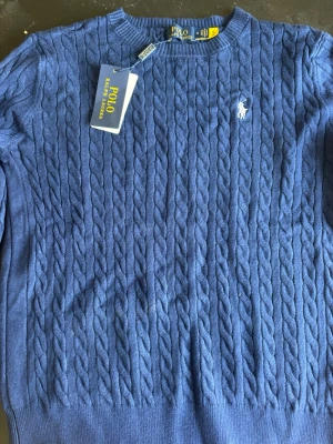 Blå kabelstickad tröja Polo Ralph Lauren - Kabelstickad blå tröja från Polo Ralph Lauren med rund halsringning och den klassiska vita logobroderingen på bröstet. Tröjan har ribbade muddar vid ärmslut och nederkant, perfekt för dig som gillar en tidlös och stilren look.