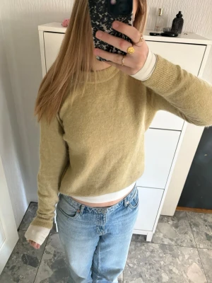 Grön/gul stickad tröja med rund hals - Mysig beige stickad tröja med rund halsringning och långa ärmar. Tröjan har ribbade muddar vid ärmslut och nederkant, och är tillverkad i ett mjukt material som ser ut att vara ullblandning. Perfekt för lager på lager och enkel att matcha med jeans.