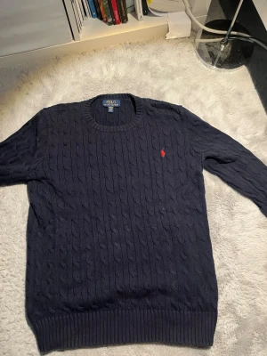 Mörkblå kabelstickad tröja Polo Ralph Lauren - Kabelstickad mörkblå tröja från Polo Ralph Lauren med rund halsringning och den klassiska röda logotypen broderad på bröstet. Tröjan har ribbade muddar vid ärmslut och nederkant. Perfekt för dig som gillar stilrena och tidlösa plagg. 8/10 skick