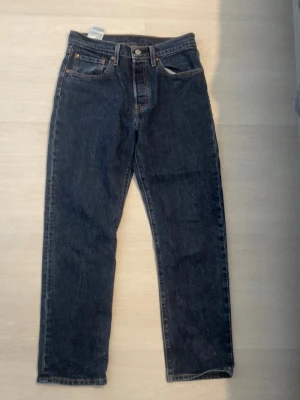 Levi's 501 blå raka jeans - Klassiska Levi's 501 jeans i mörkblå denim med raka ben och hög midja. Femficksmodell med knappgylf och snygga kontrastsömmar. Perfekta för en avslappnad och tidlös stil. Ikonisk läderpatch bak i midjan.