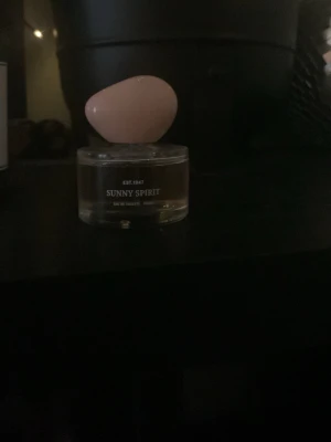 Sunny Spirit parfym med rosa kork - En parfym från Sunny Spirit med en rund, transparent glasflaska och en unik, oval rosa kork. Flaskan har en modern och stilren design som passar perfekt på sminkbordet. Doften är Eau de Parfum och flaskan har en elegant etikett.