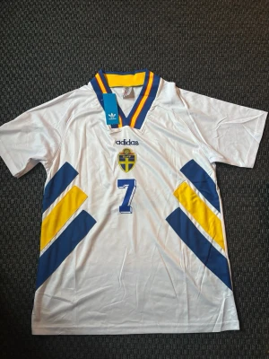 Svensk landslagströja Adidas nr 7 - Säljer en vit svensk landslagströja från Adidas med nummer 7 på bröstet. Tröjan har blå och gula detaljer, SVFF-emblem och en krage med blå, gul och röd rand. Perfekt för dig som älskar fotboll och vill stötta Sverige.