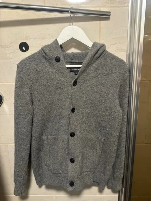 Grå luvkofta med knappar - Mysig grå kofta med luva och knäppning framtill. Tillverkad i ett mjukt, stickat material och har två fickor framtill. Perfekt för kyliga dagar och enkel att matcha med olika outfits. Passar 170-175