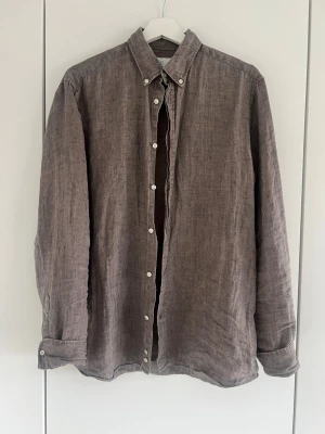 Brun linneskjorta från Bläck - Snygg brun skjorta från Bläck i linne med klassisk krage och vita knappar. Skjortan har lång ärm och en avslappnad men ändå skräddarsydd passform. Perfekt för dig som gillar stilrena och enkla plagg.
