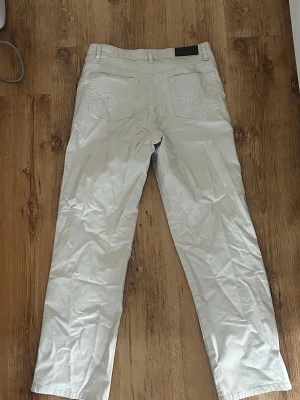 Ljusbeiga raka jeans med strass - Säljer ett par ljusbeiga jeans med raka ben och dekorativa strassdetaljer på bakfickorna. Klassisk femficksmodell med knapp och dragkedja framtill. Perfekta för dig som vill ha en clean look med lite extra bling. (vintage)