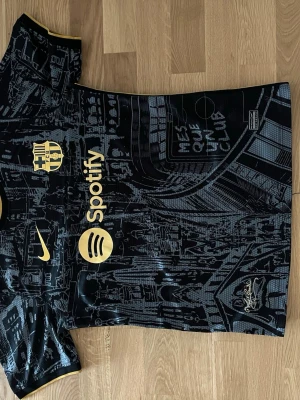 Svart Barcelona matchtröja Nike - Snygg svart Barcelona matchtröja med guldfärgade detaljer och tryck från Nike. Tröjan har stadsmotiv i grått, klubbmärke och sponsorloggor på bröstet samt texten 'Més que un club'. Perfekt för dig som älskar fotboll och vill sticka ut på planen.