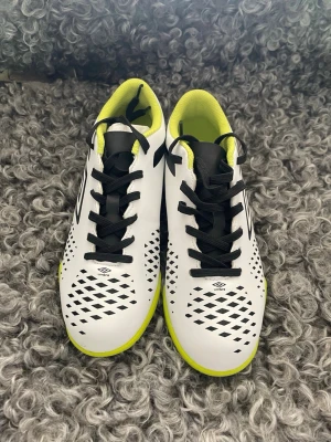 Vita Umbro fotbollsskor med neon - Snygga vita fotbollsskor från Umbro med svart logga och detaljer, samt neongul innersula och sula. Skorna har svart snörning och ett mönster av svarta rutor på ovandelen. Perfekta för dig som vill sticka ut på planen!
