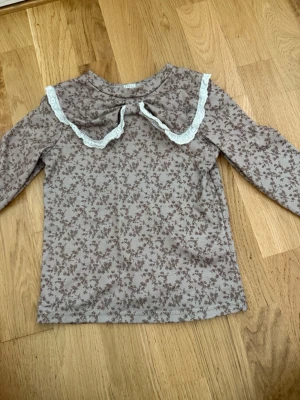 Beige blommig topp med spets från SHEIN - Söt beige långärmad topp från SHEIN med små bruna blommor och en stor krage dekorerad med vit spets. Perfekt för dig som gillar romantiska detaljer och mjuka färger.