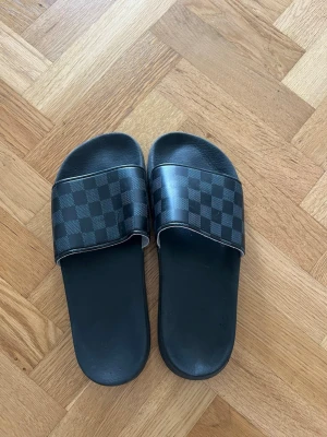 Svarta slides med rutigt mönster - Säljer ett par svarta slides med bred rem i syntetmaterial och ett stilrent rutigt mönster i grått och svart. Klassisk slip-in modell med platt sula, perfekt för chill dagar. Märket är ej synligt men designen är clean och modern.