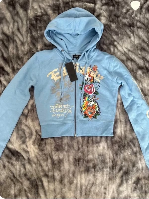 ed hardy tracksuit - Säljer en ljusblå hoodie från Ed Hardy med dragkedja och stora färgglada tatueringsinspirerade tryck fram och bak. Hoodien har huva, långa ärmar och coola detaljer som dödskallar, rosor och text. Perfekt för dig som gillar streetwear och statement-plagg.