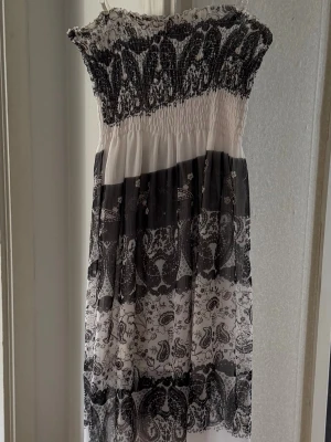 Svartvit långklänning med mönster - Säljer en svartvit långklänning med smock upptill och axelbandslös design. Klänningen har ett detaljerat paisley- och blommönster i olika sektioner och är tillverkad i ett lätt, luftigt material som passar perfekt till sommaren.