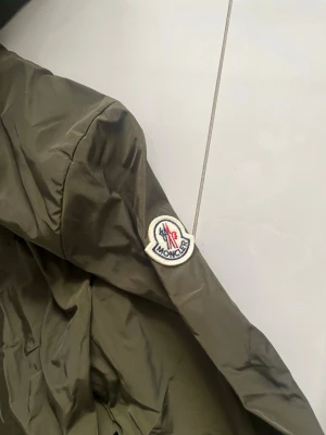 Olivgrön vindjacka från Moncler - Snygg olivgrön vindjacka från Moncler med huva och dragkedja framtill. Jackan har Moncler-logga på ärmen och är tillverkad i ett lätt, glansigt material som skyddar mot vind. Perfekt för dig som vill ha en stilren och exklusiv look.
