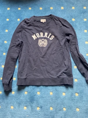 Mörkblå sweatshirt från Morris - Mörkblå sweatshirt från Morris med vit text och emblem på bröstet. Klassisk rund halsringning och långa ärmar. Tillverkad i mjukt bomullsmaterial, perfekt för en avslappnad och stilren look. Storlek S och är i bra skick 
