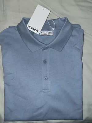 Ljusblå piké t-shirt från Name It - Snygg ljusblå piké t-shirt från Name It med klassisk krage och knappar framtill. Tillverkad i mjuk bomull och har en normal passform. Perfekt för en stilren och enkel look.